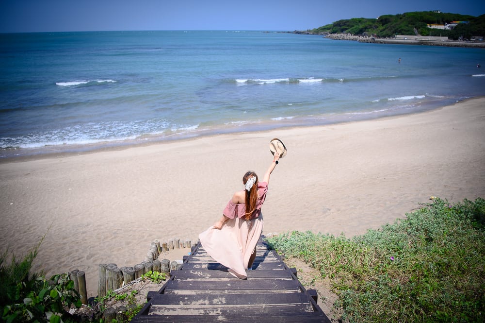 新北麟山鼻步道｜女性背對鏡頭，海邊，木棧道，沙灘，藍色海洋，草地，遠方海岸線，單手舉起帽子，裙襬隨風飄動。