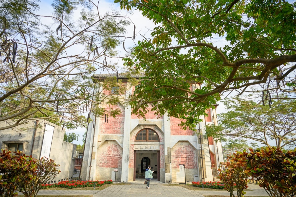 台南水道博物館｜紅磚建築、拱形門窗、行人、綠樹、枝幹、石板路、花圃。廣場中立著一棟紅磚與白色立柱交錯的高聳建築，前方