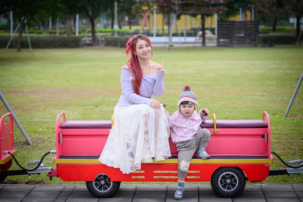 台南水道博物館｜公園、草地、婦人、小孩、紅色小火車車廂。紅髮婦人穿紫衣白裙，戴紅髮帶，端坐車廂上；小孩戴毛帽穿粉衣，