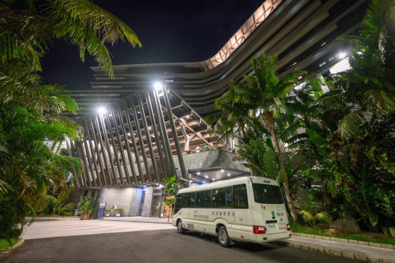 宜蘭凱渡廣場酒店 THE ARCHIPELAGO 完整指南|水上設施與熱帶園區玩法解析 16 宜蘭凱渡廣場酒店|夜間現代飯店建築外觀,白色的接駁巴士停在車道上,周圍環繞著熱帶棕櫚樹與茂密綠植,強烈的投射燈照亮入口