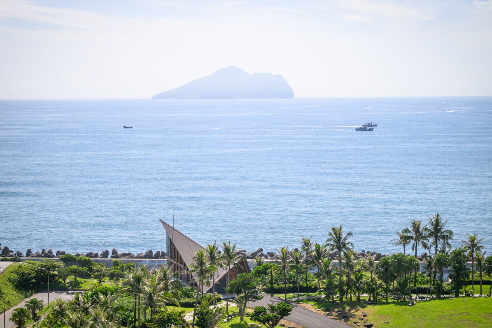 宜蘭凱渡廣場酒店｜海邊、島嶼、教堂、椰子樹、草地、船隻。遠處海平面上有孤島浮現，近景為綠地與成排椰子樹，尖頂木造建築座