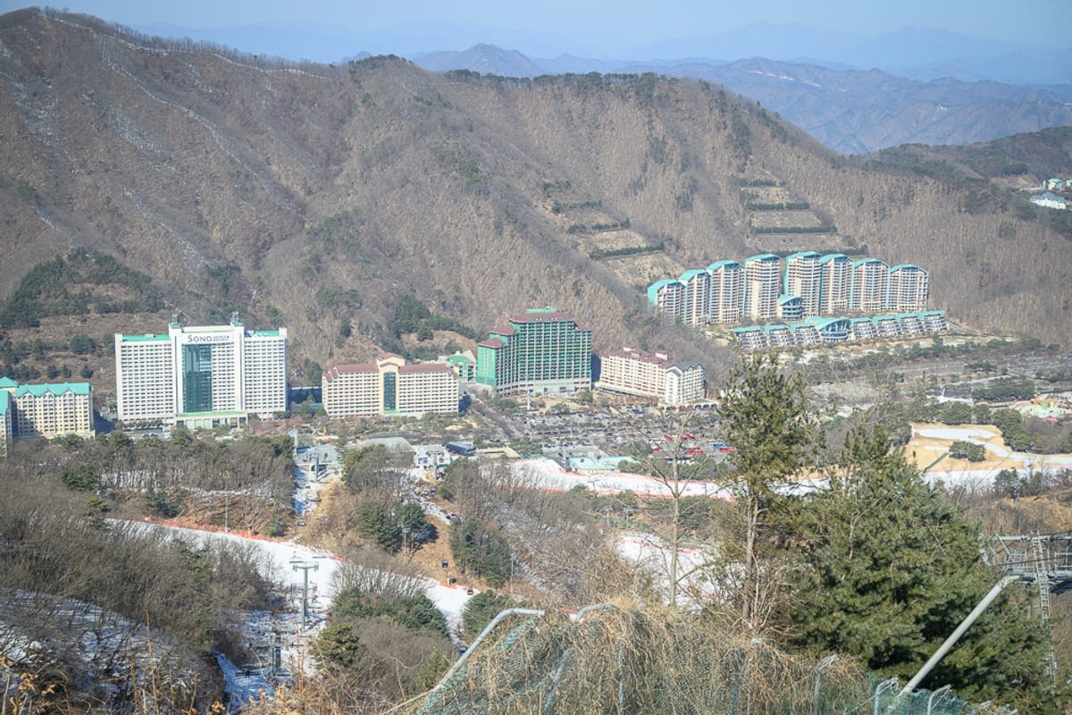 韓國首爾維瓦爾地渡假村冰雪王國｜山脈、渡假村、高樓大廈、樹林、雪道、纜車，遠山層疊，山谷間座落多棟大型飯店建築，前方鋪設零星殘雪的斜