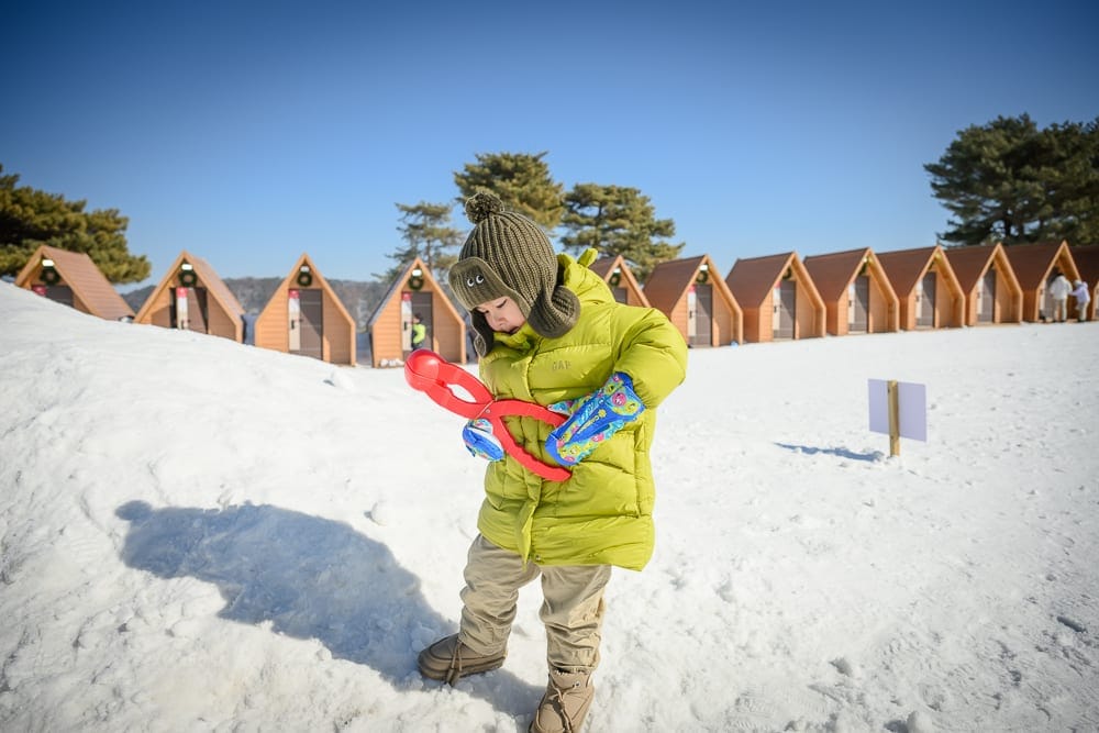 韓國首爾維瓦爾地渡假村冰雪王國｜小孩、雪地、木屋、藍天、羽絨服，白雪覆蓋的戶外場景中，一名身穿黃綠色羽絨服與毛帽的幼童站立，雙手拿著