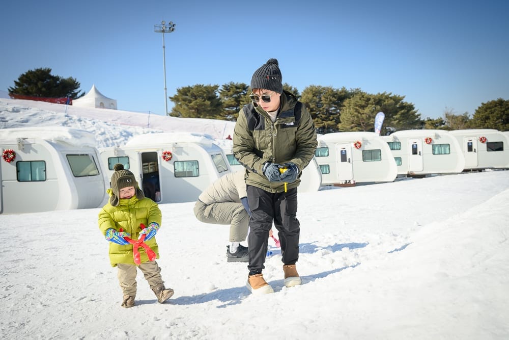 韓國首爾維瓦爾地渡假村冰雪王國｜大人、小孩、雪地、露營車、天空，戶外雪場積雪覆蓋地面，成人穿著綠色外套與毛帽站立，幼童身穿黃綠色羽絨