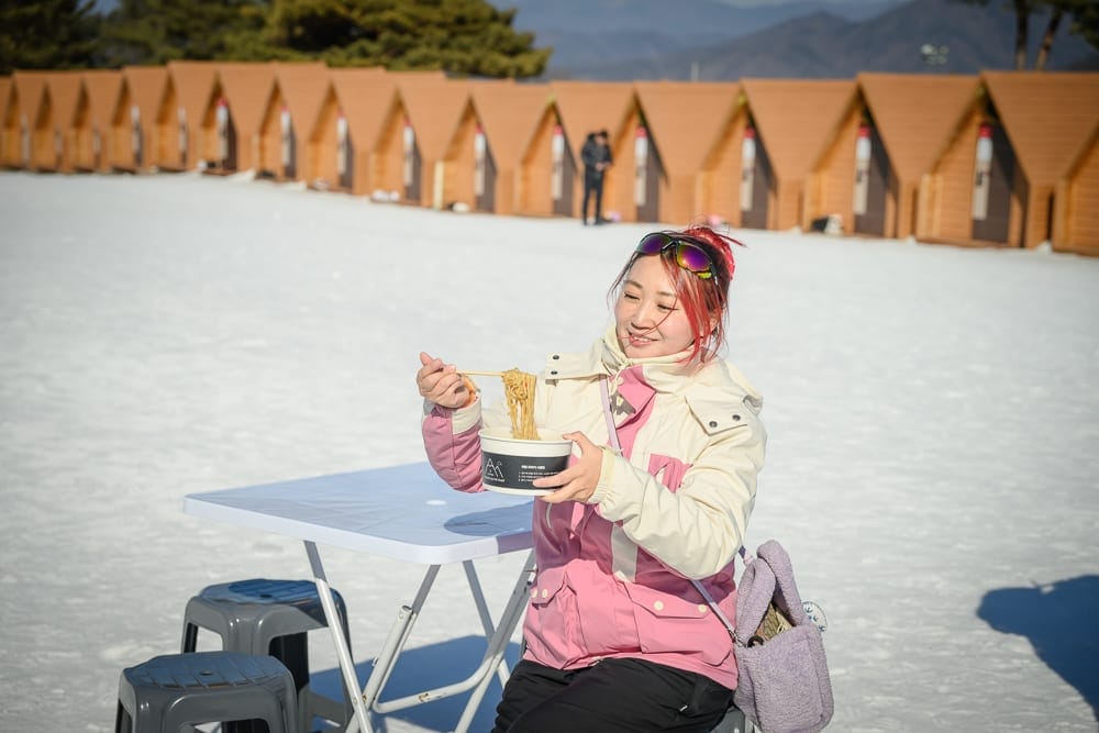 韓國首爾維瓦爾地渡假村冰雪王國｜女子，雪地，料理，桌子，小木屋，山景，女性穿著粉白相間滑雪外套與黑色長褲，手持筷子夾起碗中泡麵，坐在