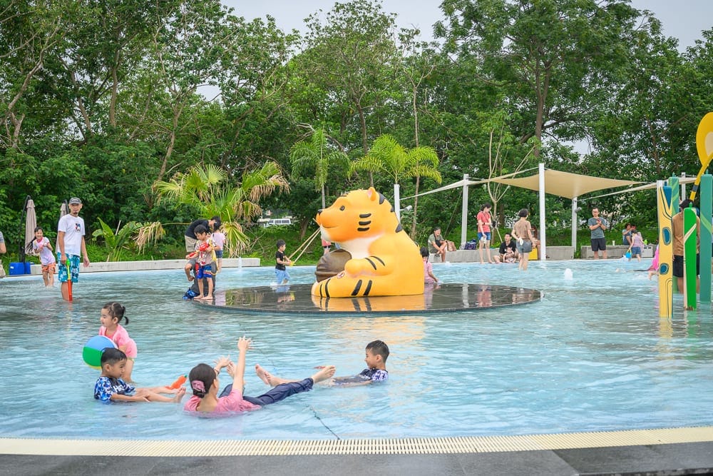 雲林虎尾632親水公園完整指南|戲水池與親子玩法解析 8 雲林虎尾632親水公園|戶外親水池、戲水池、虎型雕塑、孩童、成人、綠樹、遮陽棚、球、水槍,池中人們嬉戲,周圍站立家長。