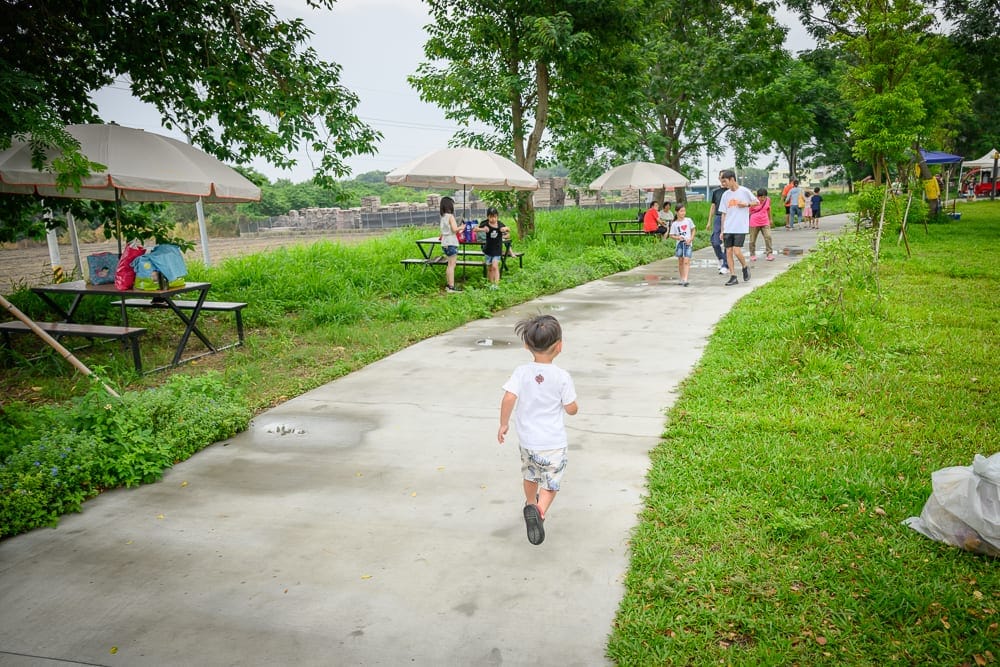 雲林虎尾632親水公園完整指南|戲水池與親子玩法解析 12 雲林虎尾632親水公園|水泥步道、草地、樹木、陽傘、野餐桌椅、孩童、行人。綠意盎然的戶外公園,穿白衣的幼童背對鏡頭向前奔跑,