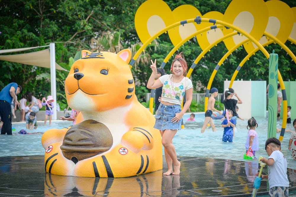 雲林虎尾632親水公園完整指南|戲水池與親子玩法解析 13 雲林虎尾632親水公園|戲水池,老虎雕像,女子,噴水設施,孩童,綠樹。女子微笑比出勝利手勢站在黃色老虎雕像旁,背景為淺水區與