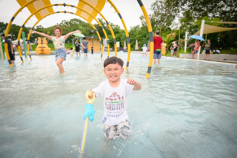 雲林虎尾632親水公園完整指南|戲水池與親子玩法解析 10 雲林虎尾632親水公園|男孩、女孩、戲水池、黃黑相間拱門、水槍、人群,戶外親水樂園裡孩童持水槍奔跑嬉戲,水花四濺,背景有綠樹