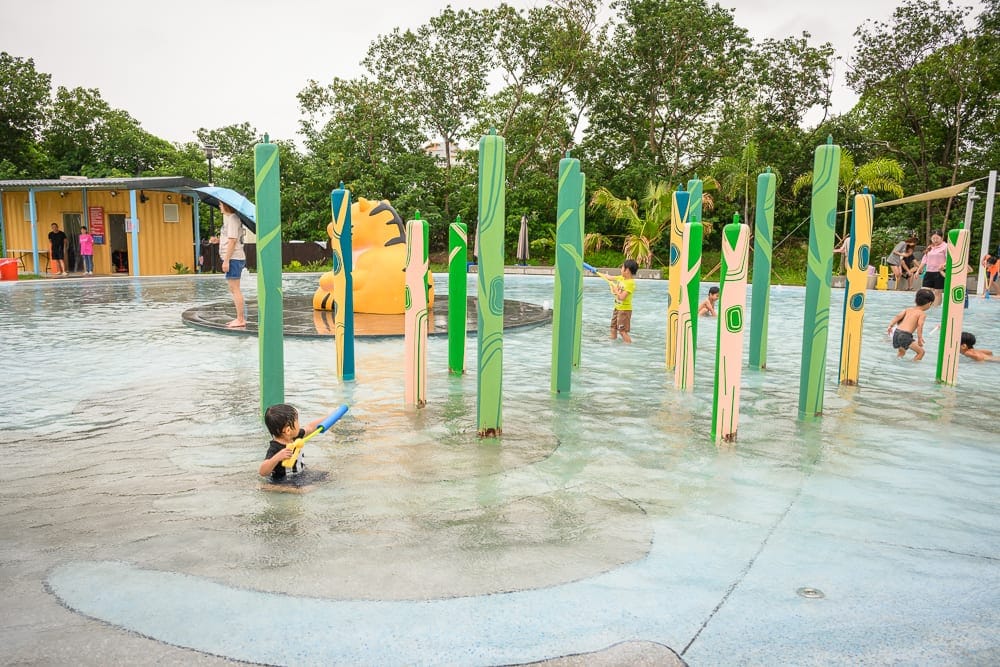 雲林虎尾632親水公園完整指南|戲水池與親子玩法解析 9 雲林虎尾632親水公園|戶外戲水池,兒童,水槍,彩色立柱,黃色虎形雕塑,黃色貨櫃屋,綠樹,陰天。多名孩童在淺水區手持水槍嬉戲