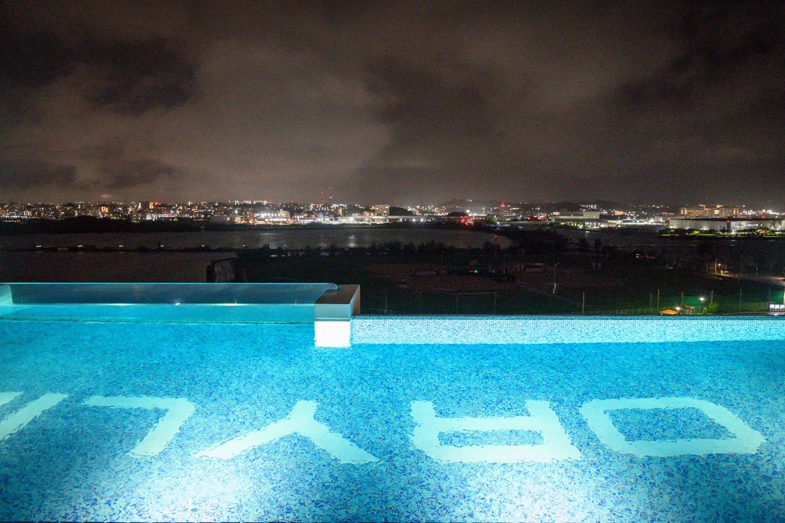 日本沖繩STORYLINE 瀬長島｜屋頂、泳池、水面、夜空、遠山、燈火，無邊際泳池底部有白色英文字母浮印，池水透出藍光，遠處為黑夜中的城