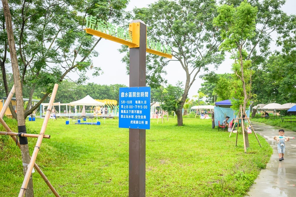 雲林虎尾632親水公園完整指南|戲水池與親子玩法解析 7 雲林虎尾632親水公園|公園、草地、指標牌、告示牌、樹木、步道、兒童。綠色草皮上有棕色柱狀指標牌,懸掛藍色告示牌,遠處有白色