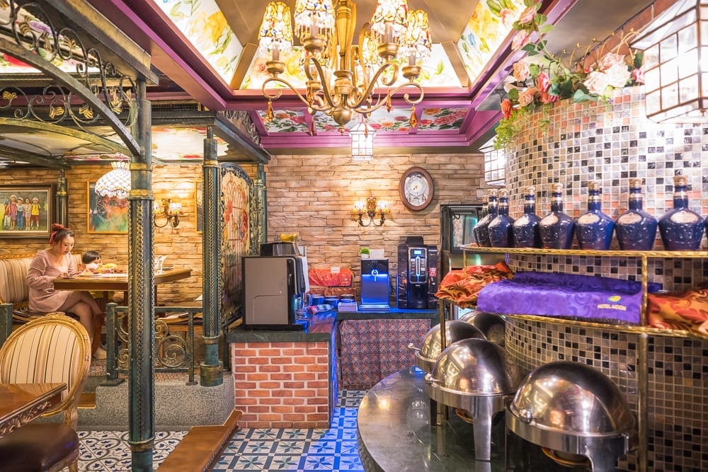 花蓮七星潭水上明月海景渡假旅店 Hotelmoon 完整攻略|歐式城堡建築與住宿體驗 12 花蓮水上明月海景旅店|室內,圓頂吊燈,石磚牆,金屬餐台,自助餐保温盆,藍瓶酒具,木製桌椅,顧客。燈光照亮陳設繁複的空間,天