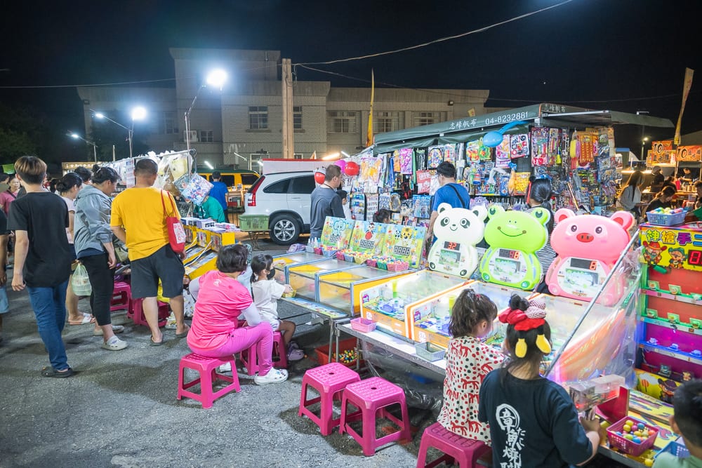 彰化秀水禮拜六夜市｜夜市，人群，彈珠台，玩具攤販，粉紅色塑膠椅，燈光照明。夜晚露天廣場擺設多個遊戲攤位，大人與孩童坐在矮