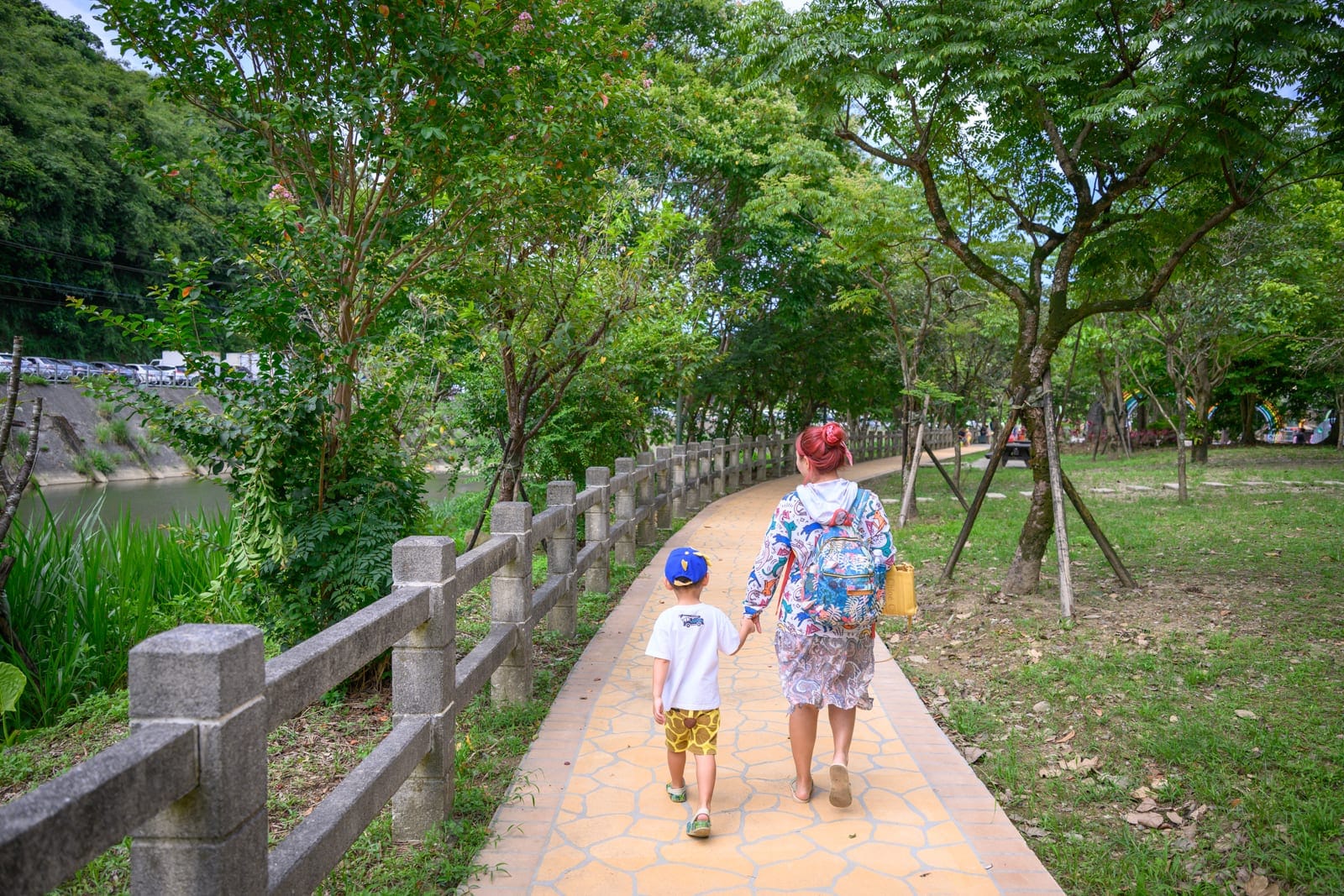 基隆暖暖親水公園完整指南|淺水戲水池與親子玩水設施解析 1 基隆暖暖親水公園|成人與戴著藍帽的孩童牽手走在鋪面步道上,右側為綠地與茂密樹木,左側為石欄杆、水溝與山壁,行人背對鏡頭