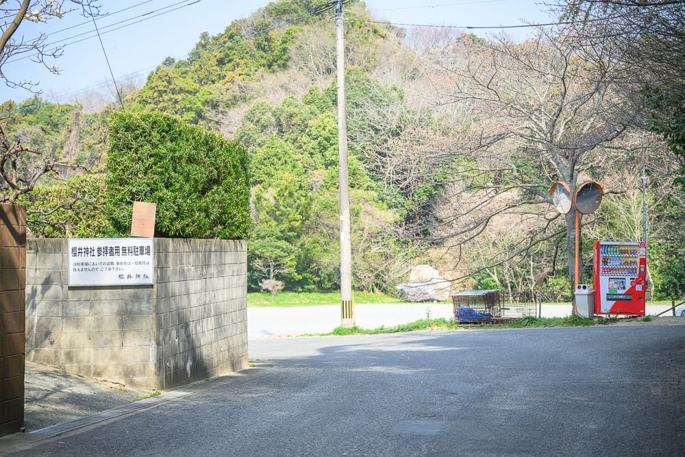 日本福岡-櫻井神社｜街道、石牆、自動販賣機、電線桿、樹林、山丘。柏油路面延伸至前方轉角，左側灰階水泥牆貼有日文告示牌，上