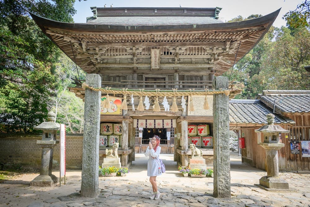 日本福岡-櫻井神社｜女子、寺廟、石柱、石燈籠、酒樽、神社、屋瓦、樹木。一名女子站在大型木造寺廟前，兩側豎立石柱與石燈籠，