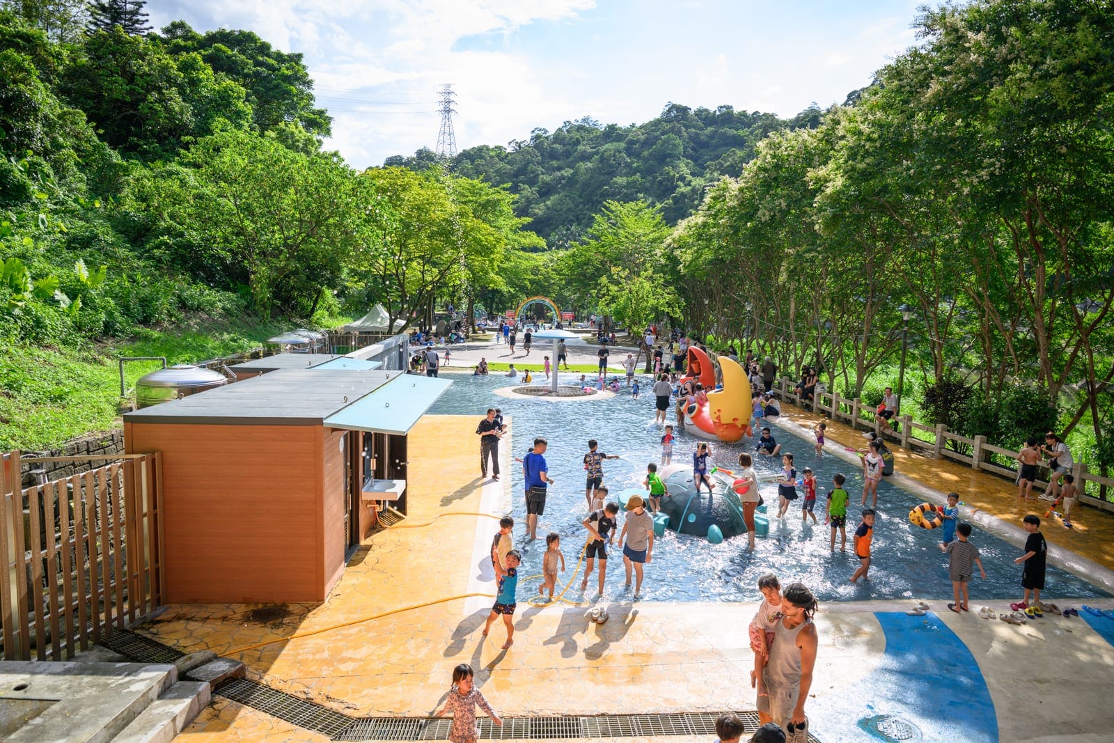 基隆暖暖親水公園完整指南|淺水戲水池與親子玩水設施解析 10 基隆暖暖親水公園|戶外水池,兒童,家長,樹林,建築,山丘,許多人在淺水區戲水與走動,四周環繞著綠樹與遠山,地面有黃色與