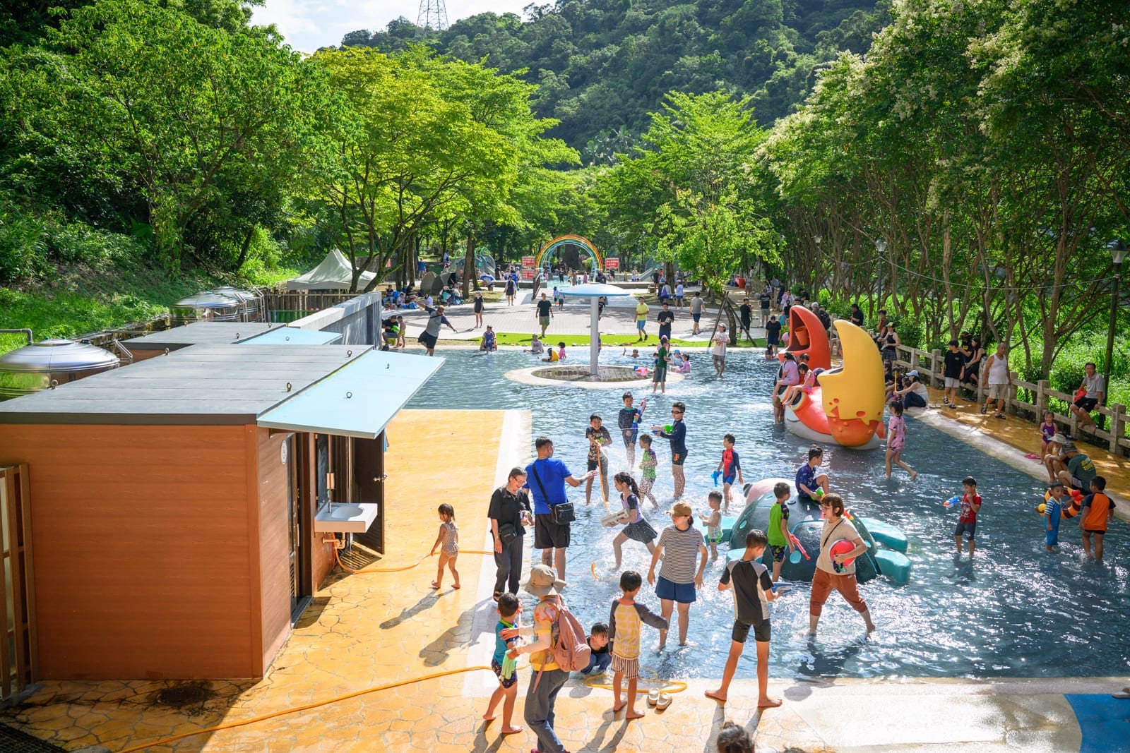 基隆暖暖親水公園完整指南|淺水戲水池與親子玩水設施解析 8 基隆暖暖親水公園|戶外戲水池,人群,兒童,水花,木屋,綠樹,山景,晴天。大人小孩在淺水中嬉戲,岸邊鋪設黃色地磚,池中立