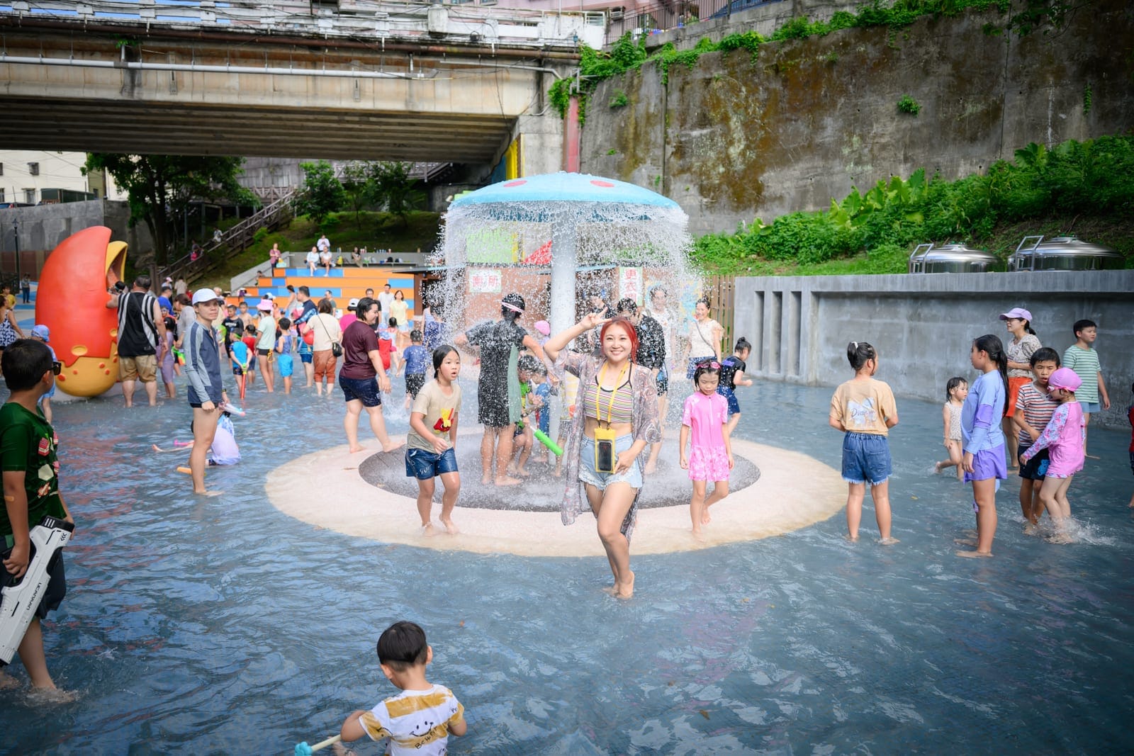 基隆暖暖親水公園完整指南|淺水戲水池與親子玩水設施解析 12 基隆暖暖親水公園|橋墩下方的戶外戲水池,大人與小孩聚集在淺水中,中央設有噴水設施,水流從蘑菇狀的頂棚落下,人們拿著水槍