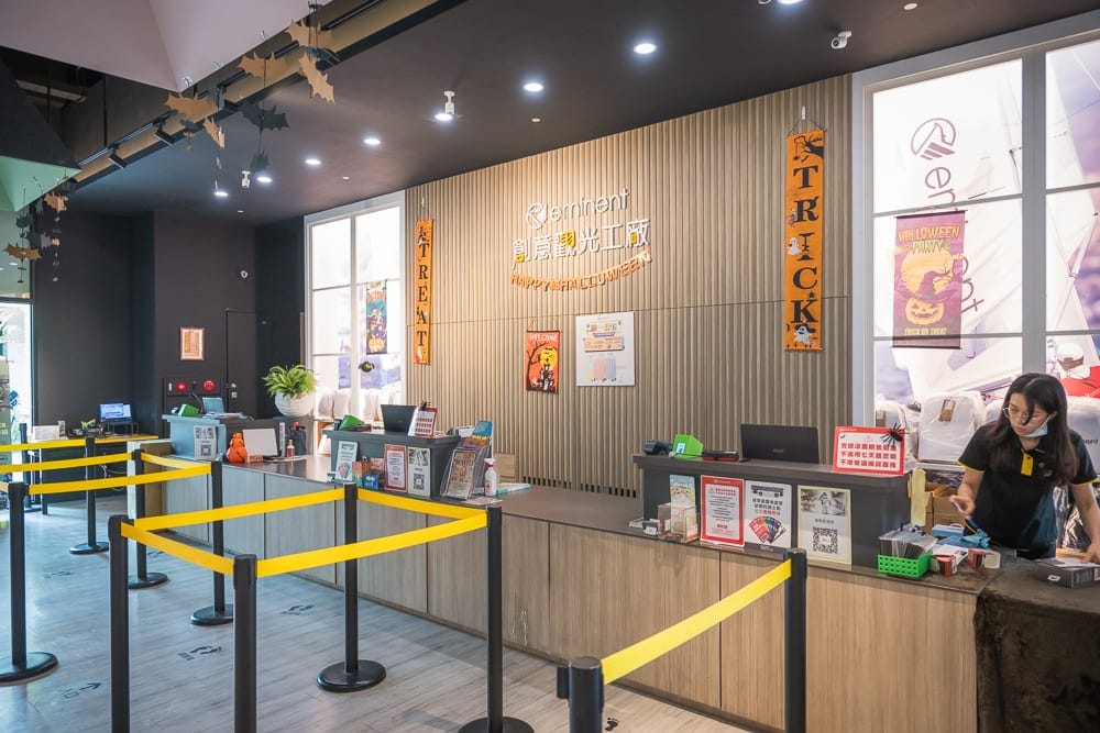 台南萬國通路創意觀光工廠|行李箱展示、親子空間與交通整理 11 台南萬國通路創意觀光工廠|室內、櫃檯、黃色警戒線、造訪時布條、工作人員。觀光工廠大廳設置木質長櫃台,前方架設黑色支柱與黃色伸縮
