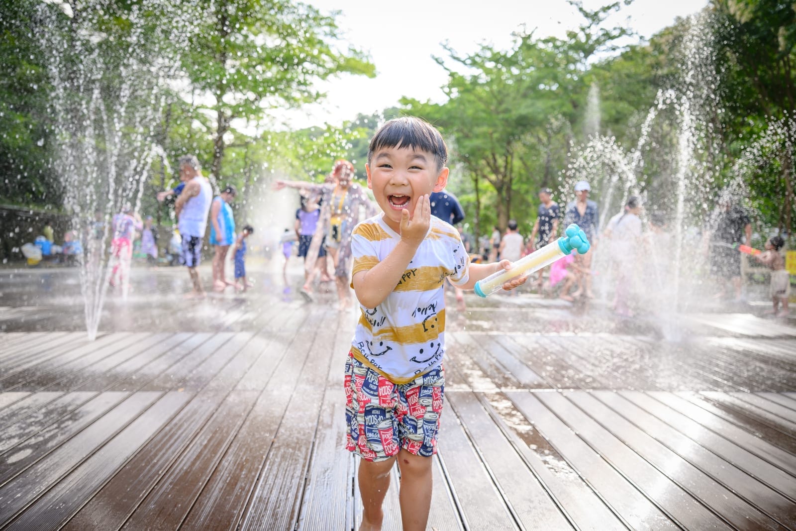 基隆暖暖親水公園完整指南|淺水戲水池與親子玩水設施解析 7 基隆暖暖親水公園|男孩,木棧道,噴水池,水槍,樹林。男孩手持水槍站在戶外木棧道上,四周噴泉水柱四濺,背景有多位行人嬉戲