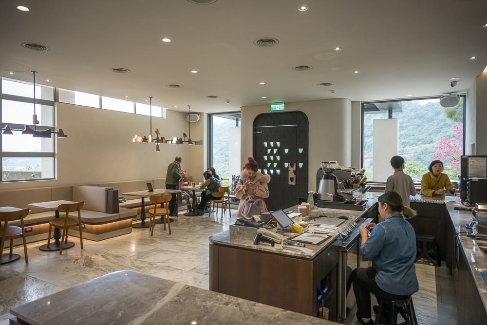 桃園福華石門水庫｜室內、咖啡廳、櫃台、木椅、沙發、客人、店員，明亮吊燈照亮座位區。靠窗處可見綠意山景。