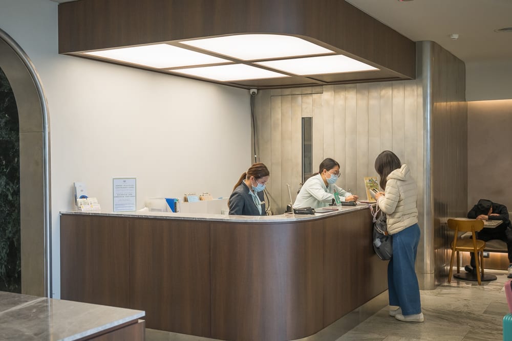 桃園福華石門水庫｜室內、櫃檯、人員、椅子、牆壁。深褐色木質櫃檯後方有兩位佩戴口罩的工作人員，櫃檯前方站著一位穿著羽絨衣