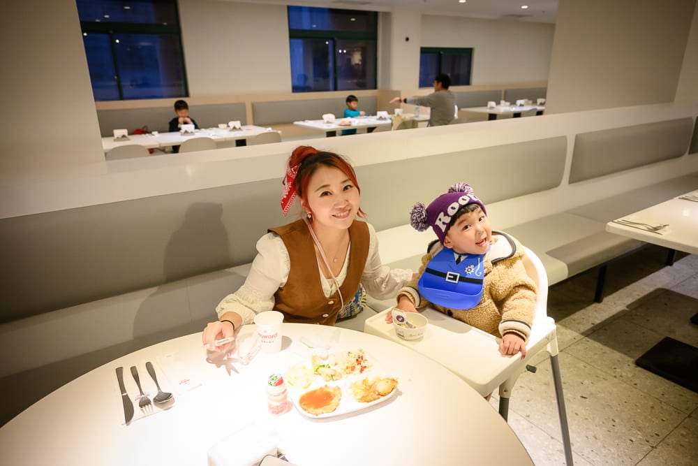 桃園福華石門水庫｜室內，餐桌，女性，小孩，兒童椅，食物，餐具，長沙發。女性與戴毛帽幼童坐在白色圓桌旁，桌上擺放盤裝食物
