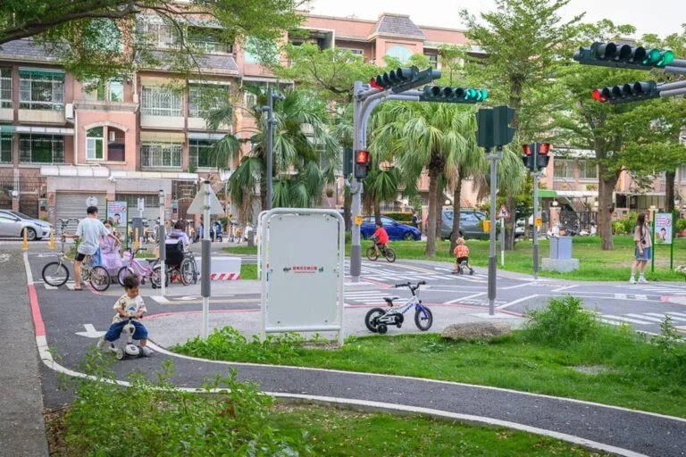 台南光賢交通公園｜戶外公園交通教育場地，兒童騎乘自行車與滑步車，路口設有小型紅綠燈、行人穿越道與標誌牌，背景為綠樹、草