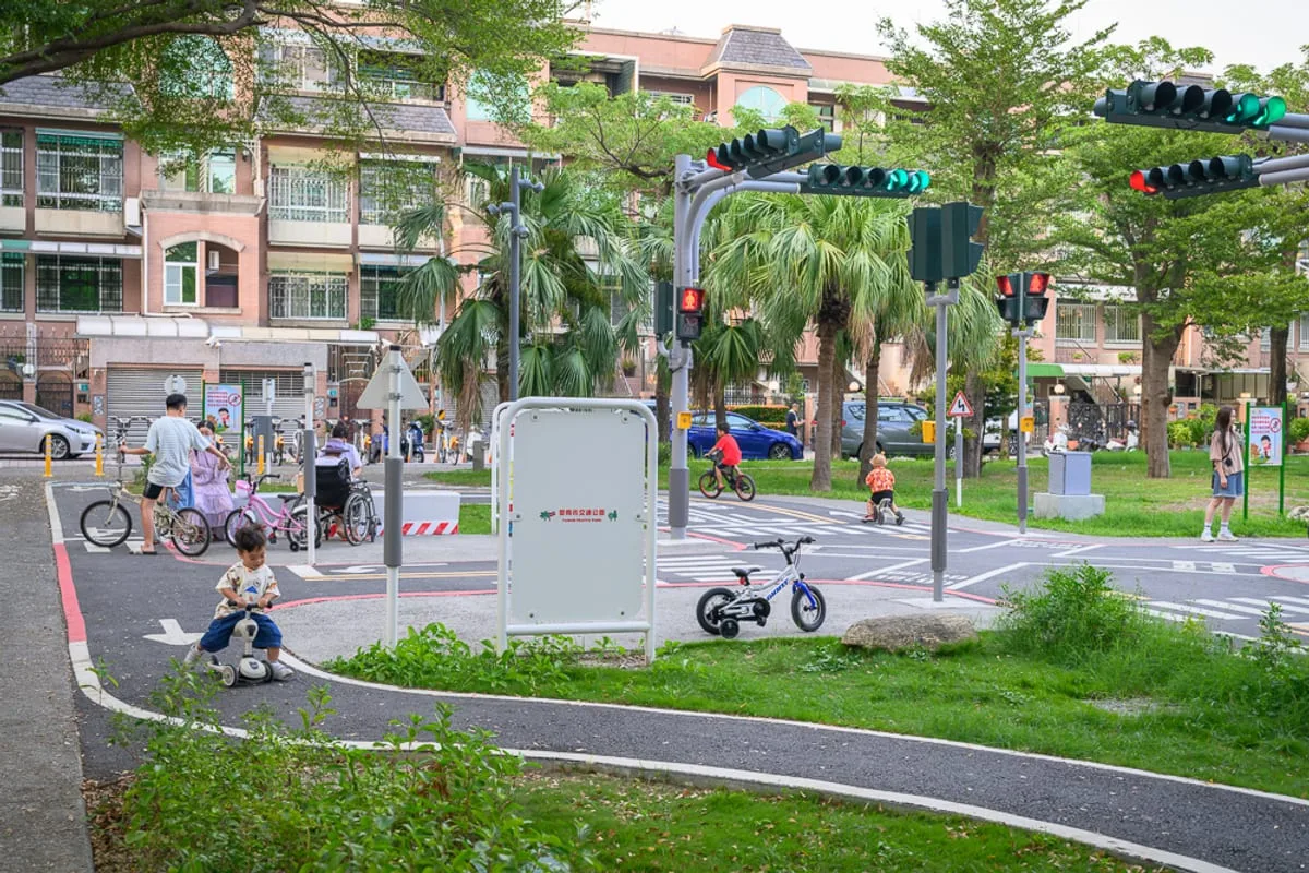 台南光賢交通公園｜戶外公園交通教育場地，兒童騎乘自行車與滑步車，路口設有小型紅綠燈、行人穿越道與標誌牌，背景為綠樹、草