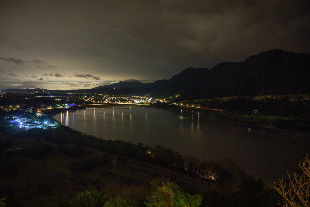 桃園福華石門水庫｜夜間高處俯瞰，河水、橋梁、山脈、遠方聚落燈火，水面反射街燈光芒，陰暗天空覆蓋山巒。