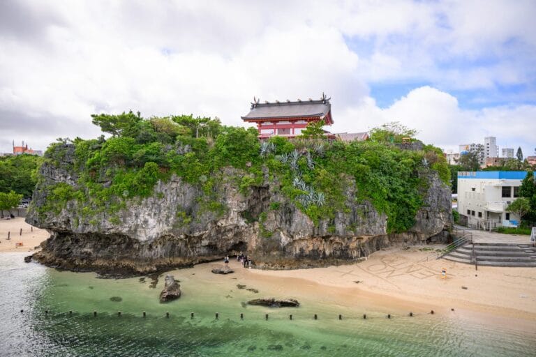 日本沖繩波上宮陸橋拍攝點與波之上海灘｜寺廟,海邊,岩石,沙灘,海水,遊客,綠樹,建築。陡峭的海蝕平台上建有紅柱白牆的日式寺廟，四周覆蓋綠樹