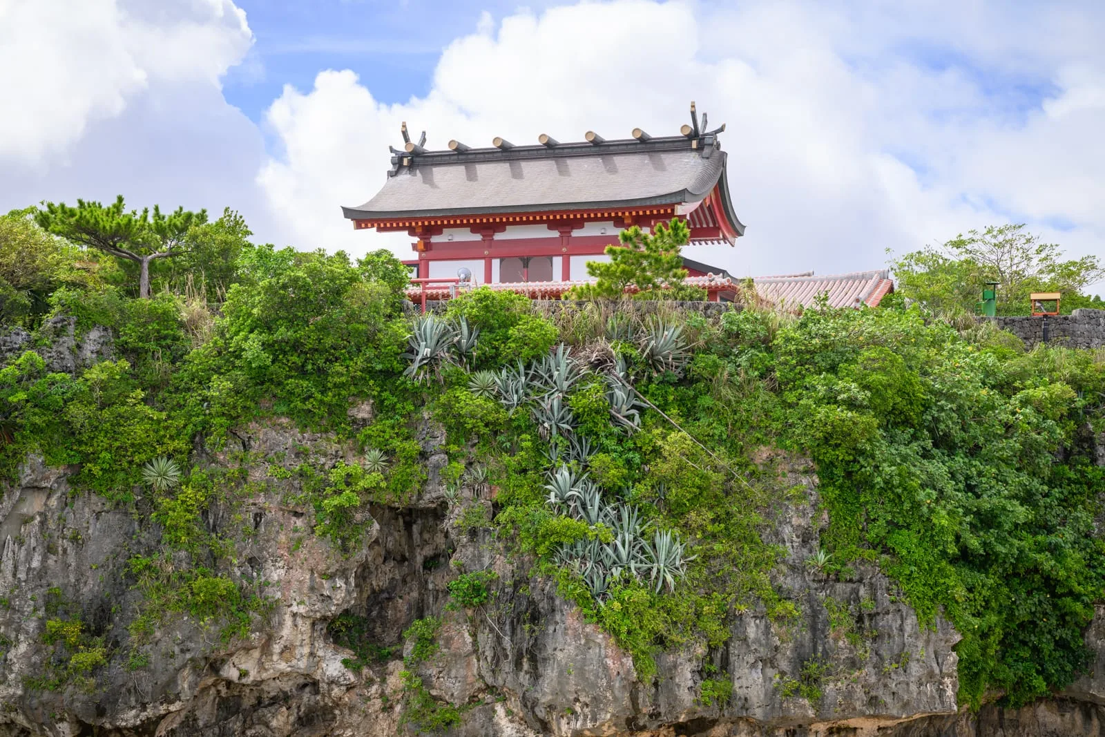 日本沖繩波上宮陸橋拍攝點與波之上海灘｜寺廟、懸崖、樹木、龍舌蘭、藍天、白雲。紅白色日式神社建築坐落於岩壁上方，峭壁長滿茂密灌木與放射狀葉片