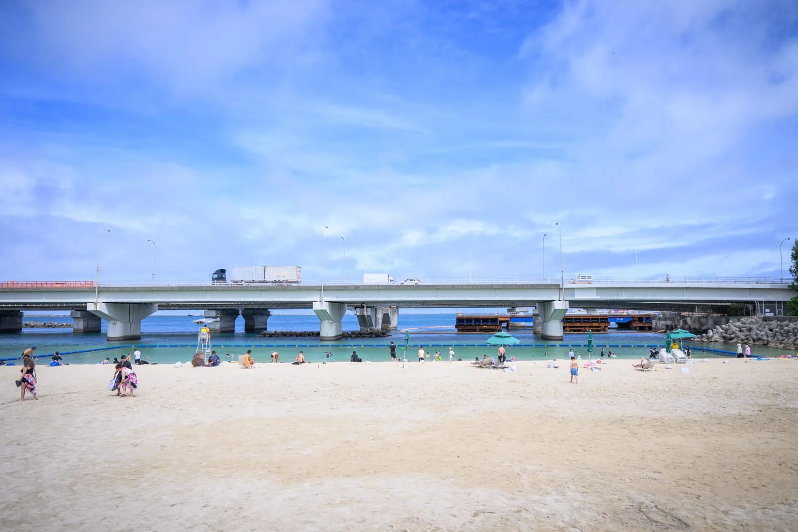 日本沖繩波上宮陸橋拍攝點與波之上海灘｜海邊，沙灘，跨海大橋，海水，人群，遮陽傘，藍天白雲。遼闊沙灘上散落著戲水遊客，遠方高架橋上有車輛行駛