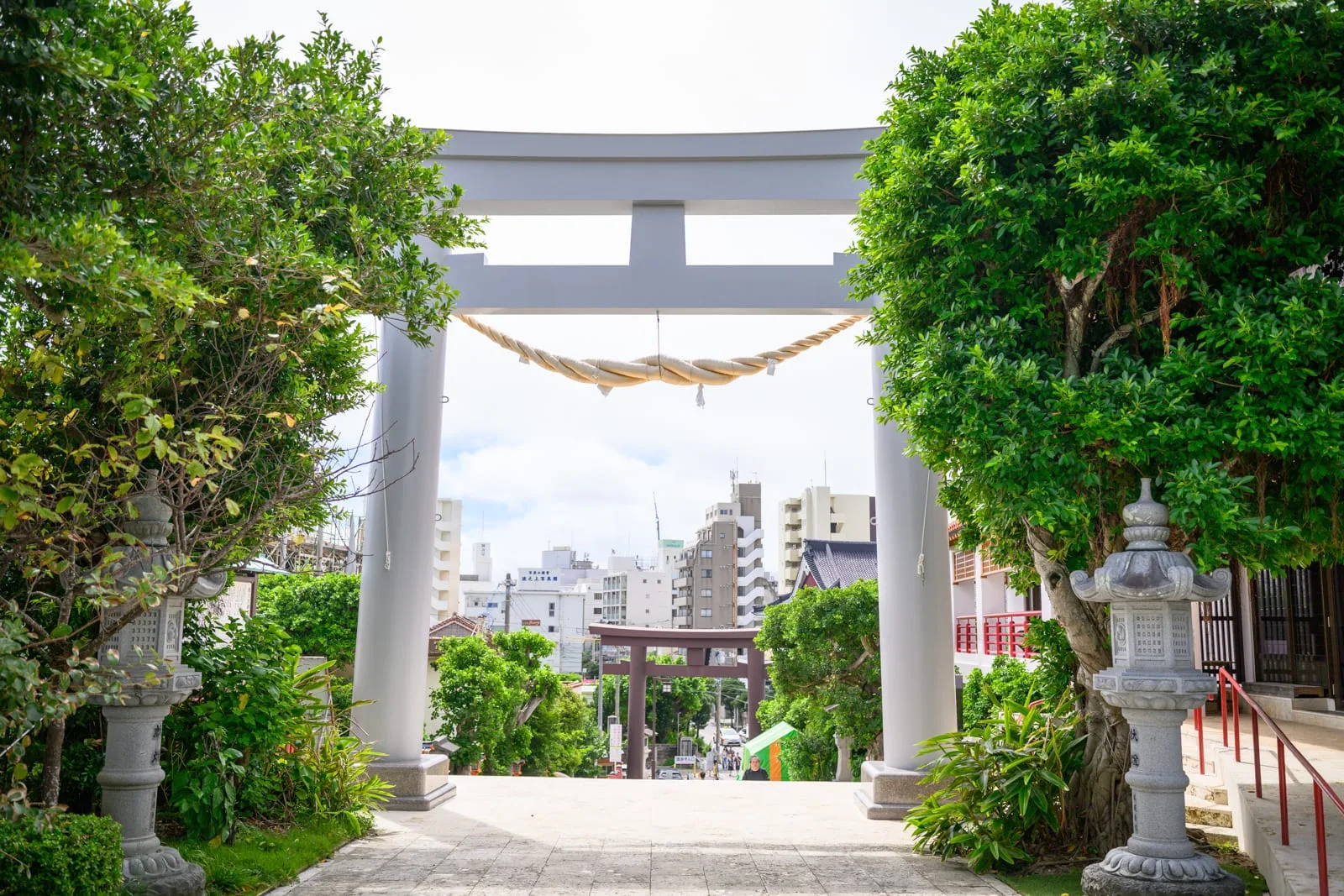 日本沖繩波上宮陸橋拍攝點與波之上海灘｜白色大鳥居、石燈籠、繩下垂、綠樹、神社入口、階梯、遠方城市大樓、晴朗天空，戶外神社場景。