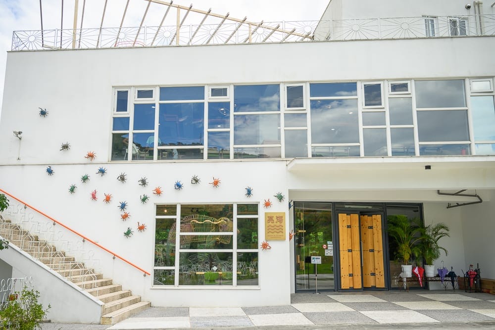 新北亮山川景觀休閒園區｜白色建築物，外牆裝飾多隻彩色螃蟹與金色小招牌，左側有戶外階梯與橘紅色扶手，一樓設有木質雙扇大門與大片