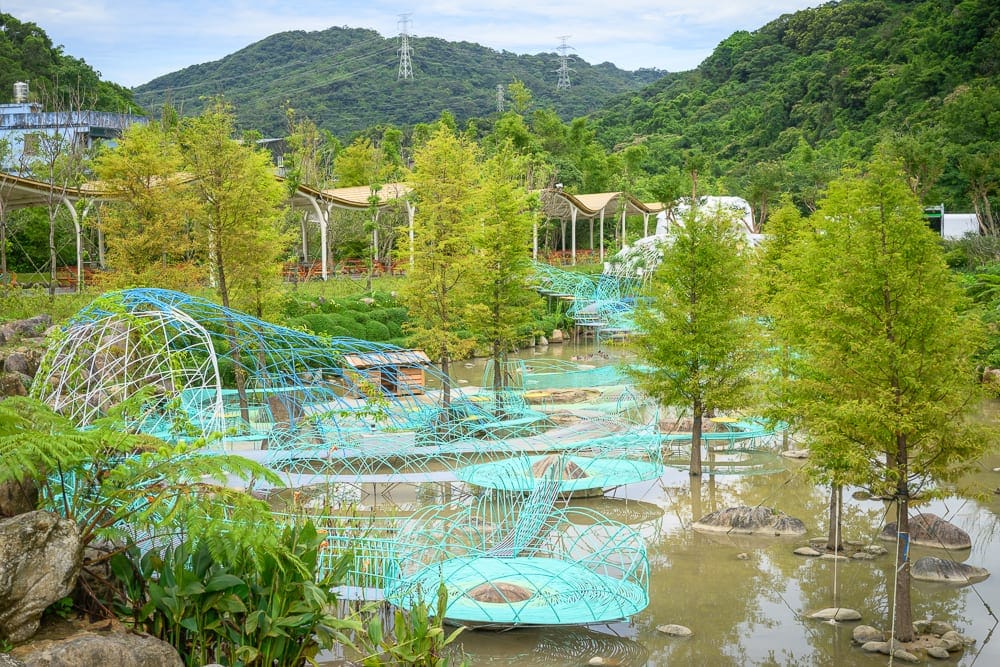 新北亮山川景觀休閒園區｜水池、落羽松、藍色金屬網狀藝術裝置、涼亭、遠山、高壓電塔。淺淺水面上錯落著幾棵細長樹木，水邊與水面皆