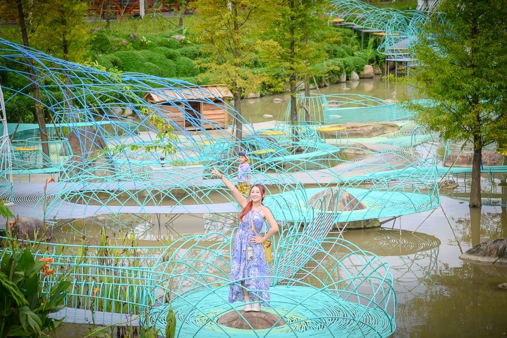 新北亮山川景觀休閒園區｜女性、藍色洋裝、竹林、水池、裝置藝術、木屋，戶外水岸景觀，藍色金屬網狀步道交織延伸，水面映照著綠樹與