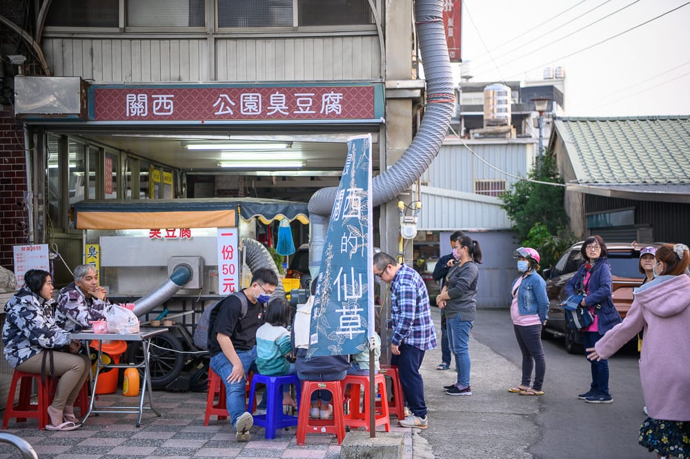 新竹關西一銀麵完整指南|黃色麵條配肉燥醬汁,號碼燈叫號候位 11 新竹關西美食|街道、招牌、攤販、人群、矮凳、臭豆腐店,騎樓下與路旁聚集多位戴口罩的顧客,店家外掛著藍色布旗,排隊人