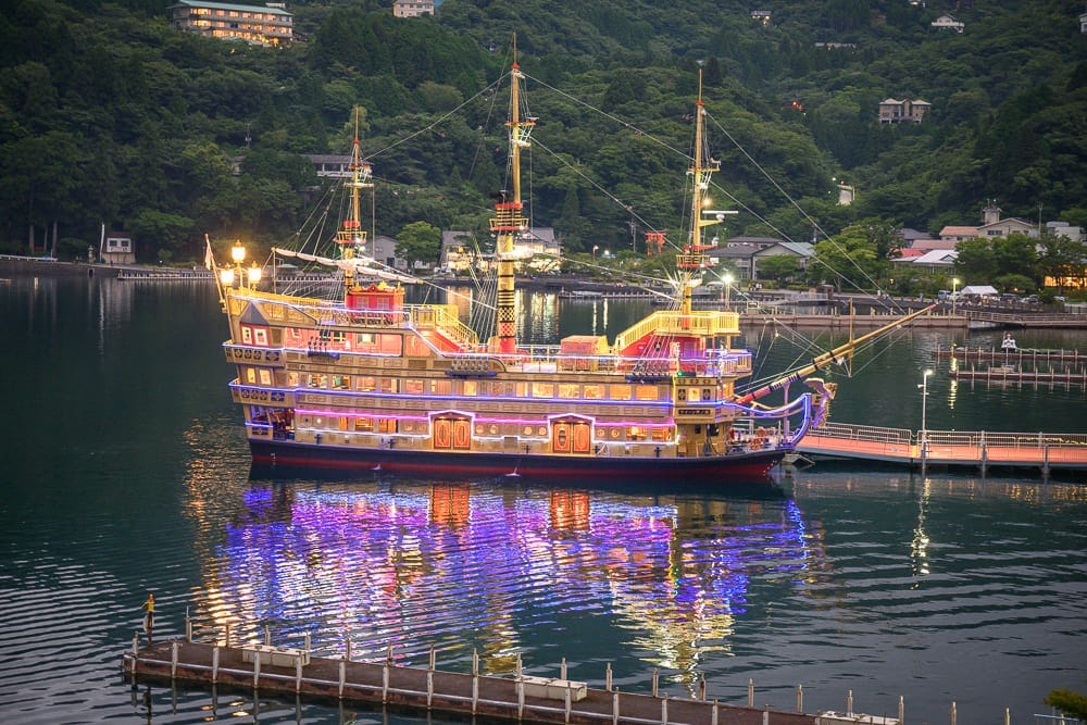 日本箱根-Rakuten STAY FUJIMI TERRACE 箱根蘆ノ湖｜船, 湖面, 碼頭, 綠山, 燈光。傍晚時分，一艘裝飾華麗的仿古帆船停泊在平靜的湖面上，船身亮起黃、