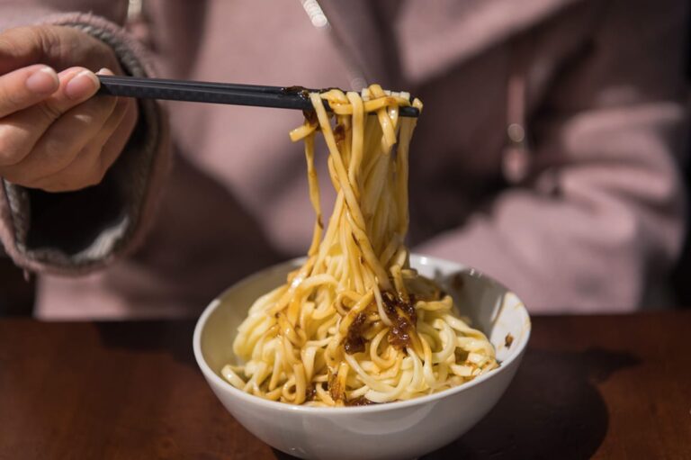 新竹關西美食｜料理，白色瓷碗，深色木桌，黑色筷子，黃色麵條，醬汁。一雙手拿著黑色筷子將白色瓷碗中的黃色麵條夾起，麵