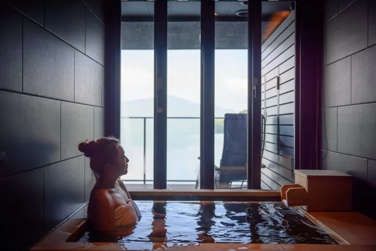 日本 Rakuten STAY FUJIMI TERRACE 箱根蘆ノ湖住宿|私人溫泉、足湯與自助入住 2 日本箱根-Rakuten STAY FUJIMI TERRACE 箱根芦ノ湖|室內、浴池、木造浴缸、女性、落地窗、陽台、山景、水面波紋。長髮紮起披著浴巾的女子浸在木造浴池中,正對