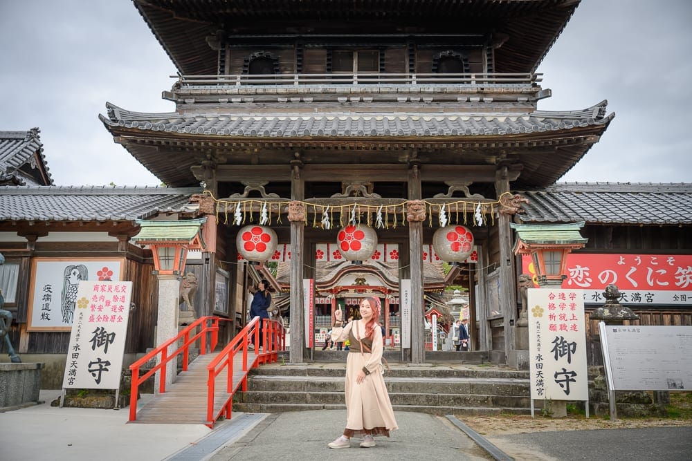 日本九州-戀木神社｜寺廟、木造建築、紅色燈籠、石階、女性，一名穿著米色連身裙的女子站在傳統木造寺廟入口前，兩側掛著圓形紅