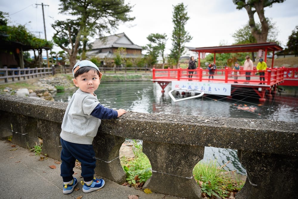 日本九州-戀木神社｜小男孩、石欄杆、池塘、紅橋、建築、樹木、遊客，戶外日間陰天。男孩戴著淺色帽子與灰藍運動服，站在石橋邊