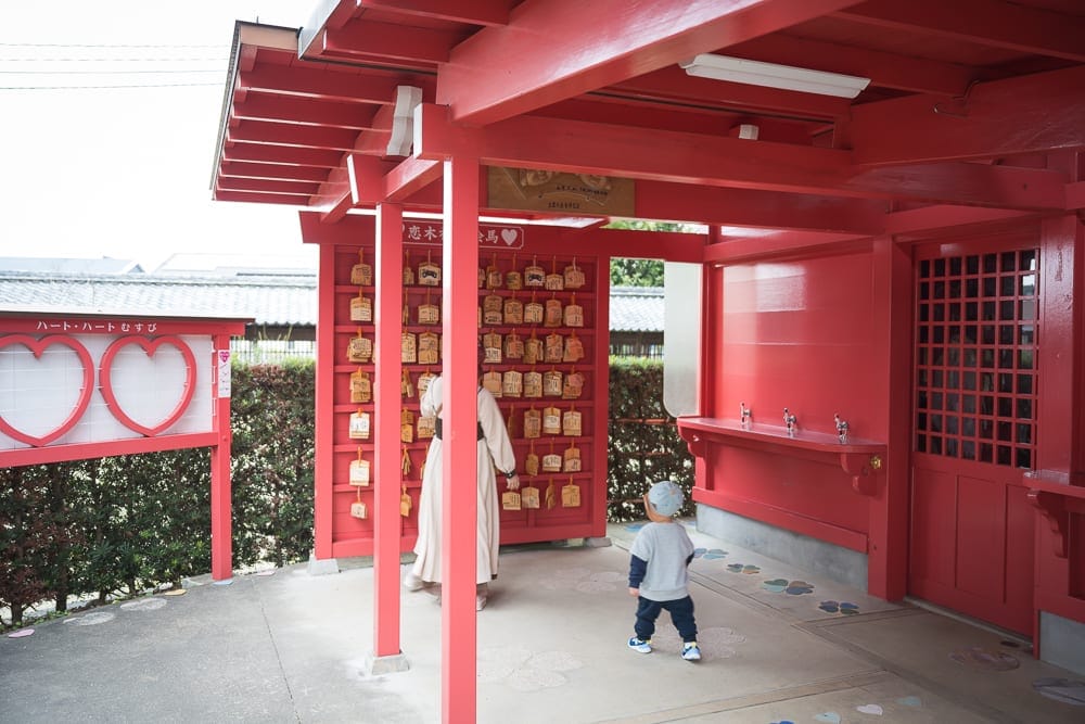 日本九州-戀木神社｜朱紅色木造神社涼亭，繪馬牆，長袖白衣女子，灰色上衣幼童，綠色樹籬，粉紅心形立牌，灰色水泥地。