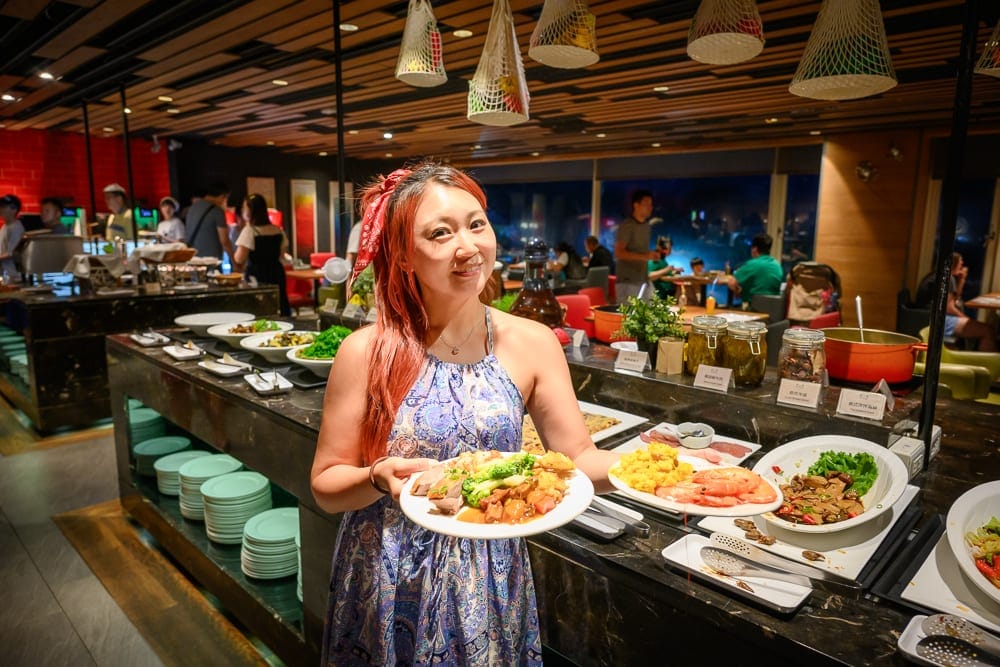 新北薆悅酒店｜室內、自助餐廳、女性、料理、盤子，染紅髮女性身穿藍色花紋洋裝，雙手各端裝滿食物的白色盤子，站在擺放多