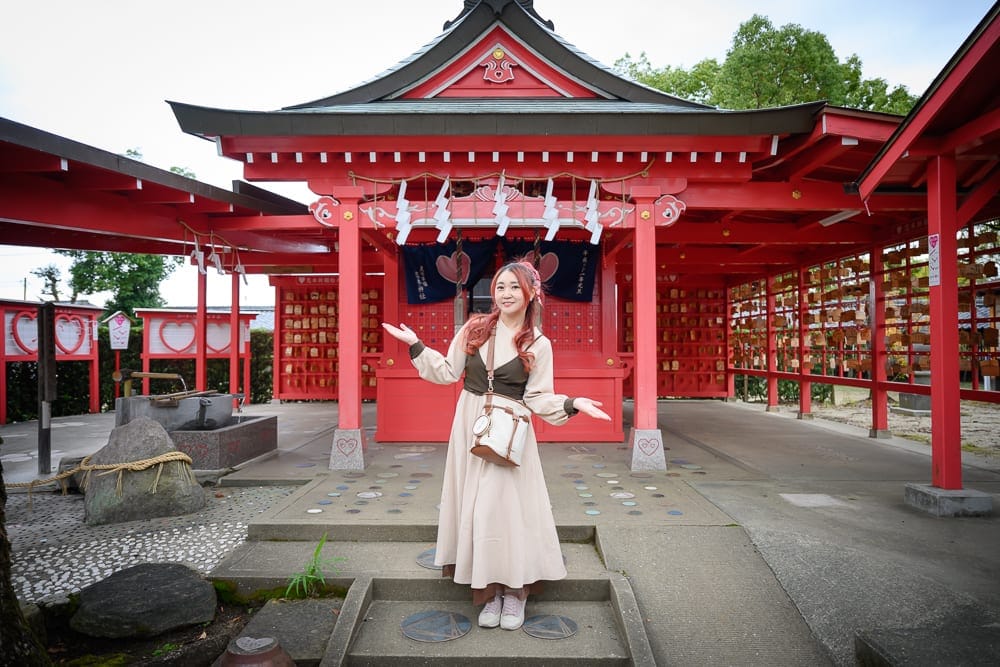日本九州-戀木神社｜寺廟、女性、紅色建築、木造迴廊、繪馬牆、石階，長髮女性身穿淺色洋裝站在鮮紅木造神社前，雙手微張，四周