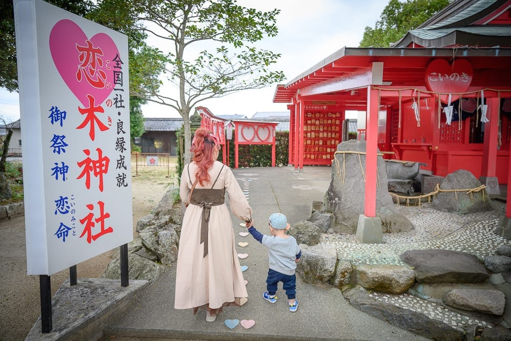 日本九州-戀木神社｜女子與孩童牽手走在神社石板路上，背對鏡頭。左側立著寫有「戀木神社」的白色大招牌，右側為紅柱建築與繫著