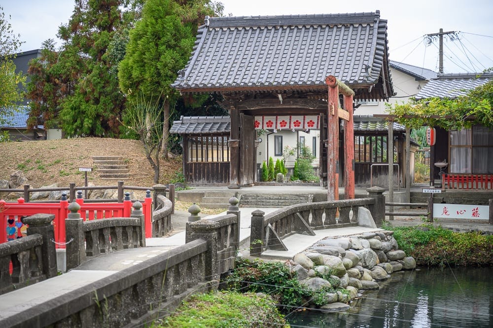 日本九州-戀木神社｜寺廟、石橋、紅欄杆橋、水池、日式瓦屋頂建築，灰白陰天光線照亮木造山門，石砌護岸與綠樹環繞池水，遠方民
